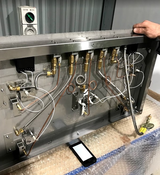 Under-bench view of commercial gas hotplate showing copper thermocouple capillary tubes with coiled strain relief, ignition generator, spark leads, and valve connections